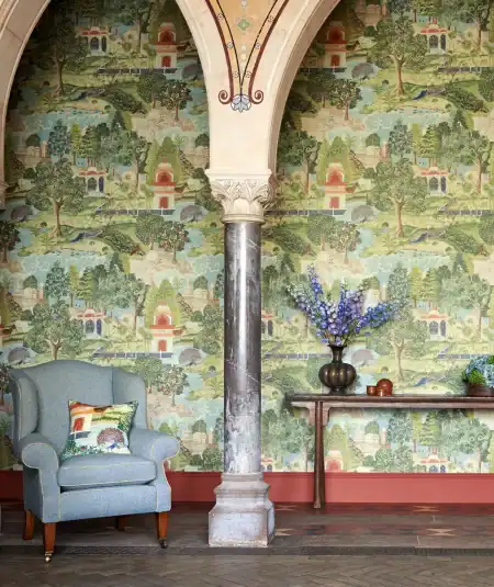 A room adorned with the Peacock Garden Green/Coral wallpaper, illustrating its transformative effect. The wallcovering features luscious fruit-laden trees, vibrant lotus flowers, and majestic peacocks, creating a sophisticated and serene Indian garden ambiance within the space.